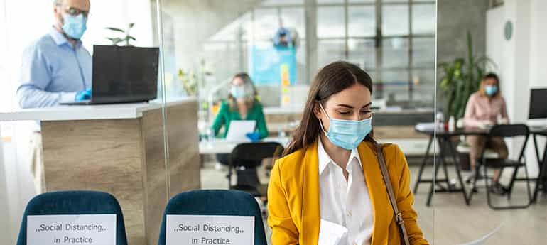 a person wearing a mask waits in the lobby outside an office space where the employees are also wearing masks